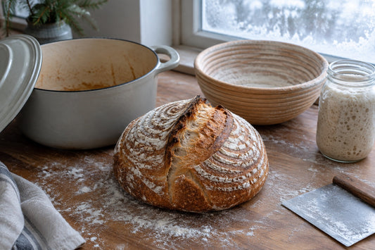 Sourdough boule beside an enamelled Dutch oven, rattan banneton and jar of active starter on a floured counter